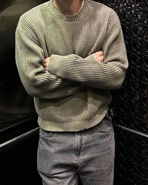 Person wearing a green sweater and gray jeans standing against a dark textured wall.
